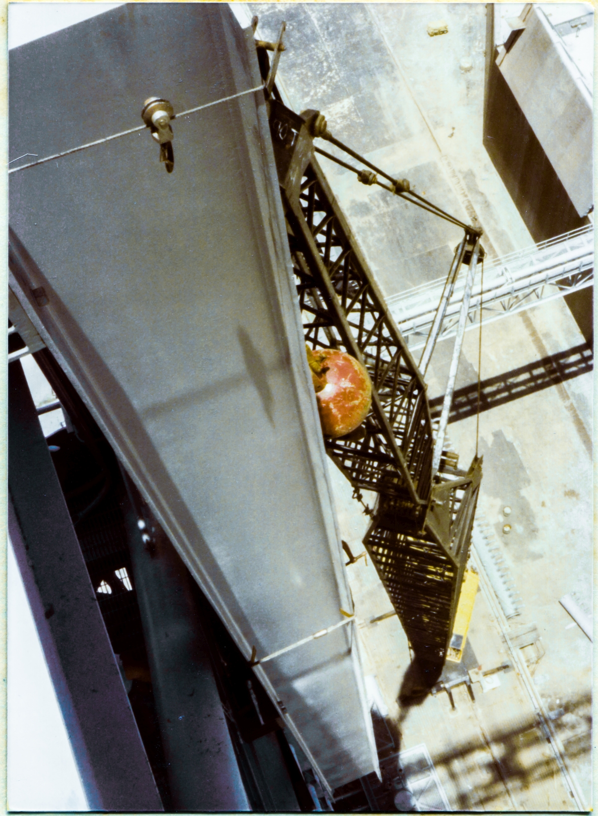 Image 085. You are on top of the Fixed Service Structure, 300 feet above the bottom of the Flame Trench at Space Shuttle Launch Complex 39-B, Kennedy Space Center, Florida, on the boom of the Hammerhead Crane, which projects outward to the east of the main body of the FSS. You are looking down the length of the web of one of the two 40-foot-long W24x104's which make up the main body of the GOX Arm Hinges Support Strongback, which is in suspension, held up by the yellow crane which you see a small part of, including the operator through his cab window, far beneath you sitting on the Pad Deck at the bottom end of a black boom which is almost 250 feet tall. High-summer midday shadows in Florida point nearly straight down beneath that which is casting them on elements of the world below. In the top right corner of the frame, the East Side Flame Deflector appears as a gray rectangle above the fifty-foot-high darkness of the East Flame Trench Wall. Immediately below it, the pale steel of the North Piping Bridge spans the Flame Trench, casting its latticework shadow on the refractory bricks that line its bottom surface. In the bottom right of the frame, the softer latticework shadow of the Hammerhead Crane, much farther above, which you are standing on, extends across the Crawlerway surface of the Pad Deck, to and beyond the invisible five-story precipice of the West Wall of the Flame Trench which is betrayed only by the very slight offset in the shadow, near the right margin of the frame. Your own shadow is in-frame, but unseeable, dwarfed into invisibility by the imagination-beggaring scale of that which you are a part of. You are, if only temporarily, part of an Ironworker's World, and it is a world apart, like no other. Anywhere. Photo by James MacLaren.